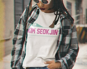 May include: A person wearing a white t-shirt with the text "I AM KIM SEOK JIN" in pink and teal. They are also wearing a black and gray plaid shirt, blue jeans, and white sunglasses. The person has dark braids.