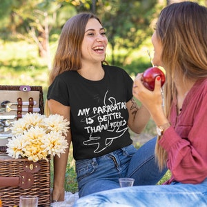 May include: Two people sitting on a blanket in a grassy area. One person is wearing a black t-shirt with the text "My Parasatal is Better Than Yours" and a white graphic of a parasail. The other person is wearing a red shirt and holding a red apple.