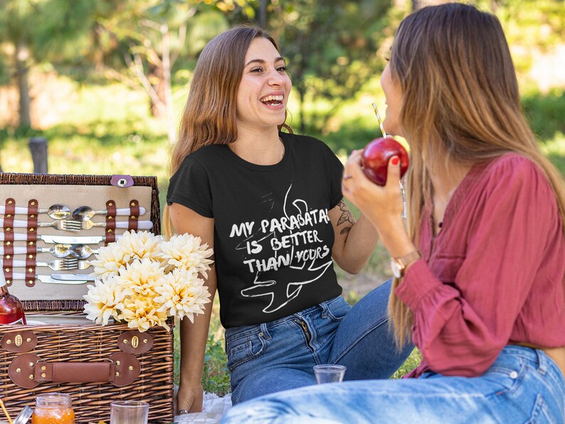May include: Two people sitting on a blanket in a grassy area. One person is wearing a black t-shirt with the text "My Parasatal is Better Than Yours" and a white graphic of a parasail. The other person is wearing a red shirt and holding a red apple.