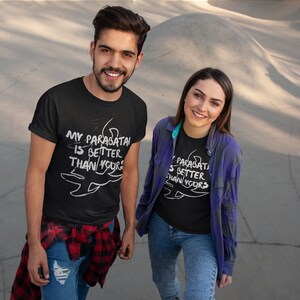 May include: Two people wearing black t-shirts with white text that reads "My parabatai is better than yours". The text is accompanied by a stylized drawing of a parabatai symbol. The people are standing in front of a concrete surface.