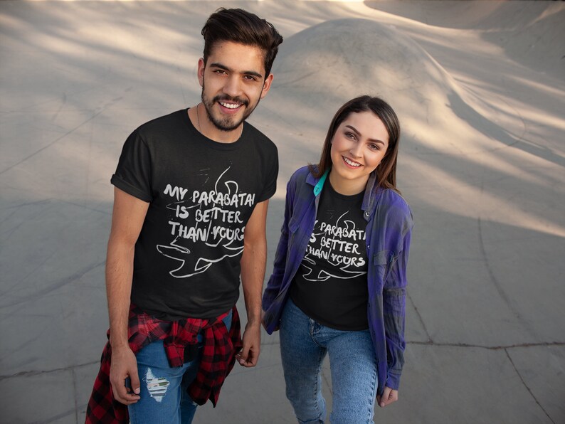 May include: Two people wearing black t-shirts with white text that reads "My parabatai is better than yours". The text is accompanied by a stylized drawing of a parabatai symbol. The people are standing in front of a concrete surface.