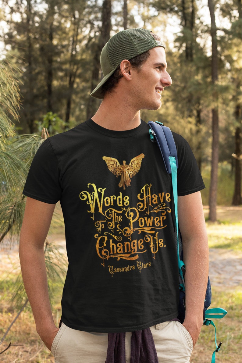 Puede incluir: Una camiseta negra con un estampado gr&aacute;fico dorado de un &aacute;ngel con alas y el texto "Words Have The Power To Change Us. Cassandra Clare".