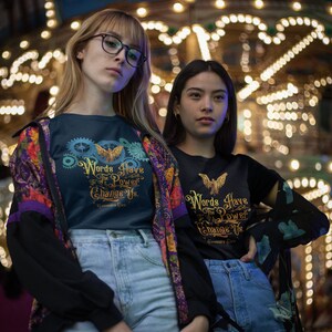 Puede incluir: Dos personas con camisetas negras con letras doradas que dicen "Words Have The Power To Change Us." Las camisetas presentan un p&aacute;jaro estilizado con las alas extendidas. Las personas est&aacute;n de pie frente a un carrusel brillantemente iluminado.