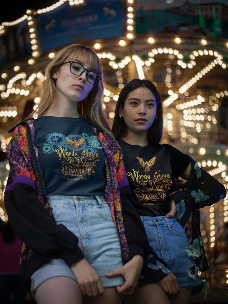 Puede incluir: Dos personas con camisetas negras con letras doradas que dicen "Words Have The Power To Change Us." Las camisetas presentan un p&aacute;jaro estilizado con las alas extendidas. Las personas est&aacute;n de pie frente a un carrusel brillantemente iluminado.