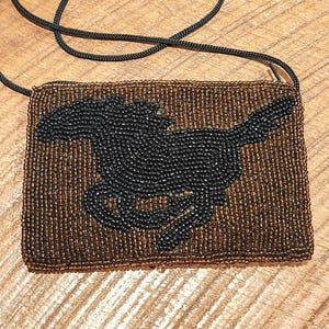 May include: A rectangular beaded crossbody bag featuring a brown background with a black horse silhouette in the center. The bag is constructed with small, uniform beads and includes a thin, black cord strap for carrying over the shoulder.