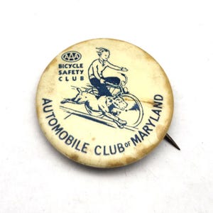 May include: Vintage Bicycle Safety Club pin from the Automobile Club of Maryland. The pin is round, with a cream-colored background and blue graphics. It features a person riding a bicycle with a dog running alongside. Text includes "Bicycle Safety Club" and "Automobile Club of Maryland."