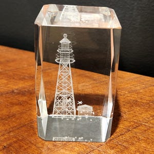 May include: A clear, rectangular crystal block with a detailed etched image of a lighthouse. The lighthouse is tall and slender, with a detailed lattice structure. A small building is etched at the base. The crystal block sits on a wooden surface.