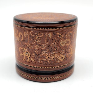 May include: A cylindrical, decorative box with a lid. The box is primarily brown with intricate gold-colored designs and black accents. The design features swirling patterns and stylized figures. The box is likely used for storage or display.