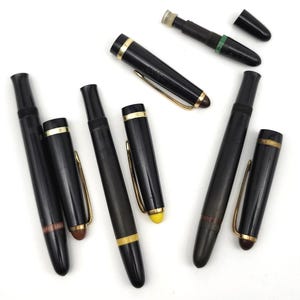 May include: A collection of black fountain pens with gold-colored accents and clips. Some pens have their caps off, revealing the nibs. The pens are arranged on a white surface, showcasing their sleek design.