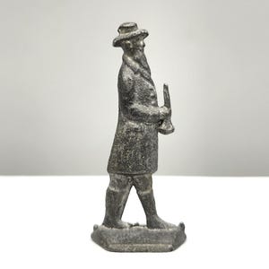 May include: A small, antique metal figurine of a man in a long coat and hat, holding a trumpet-like instrument. The figure is detailed with a textured surface and stands on a rectangular base. The overall color is a muted gray.