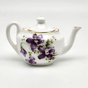 May include: A small, white ceramic teapot with a lid, handle, and spout. The teapot is decorated with purple floral designs and green leaves. The rim and spout are trimmed with gold. A charming piece for tea lovers.
