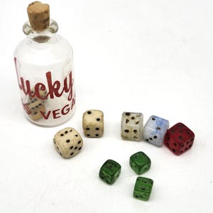 May include: A clear glass bottle with a cork stopper and the words "Lucky Vegas" in red script. Several dice in various colors, including cream, green, and red, are scattered around the bottle.