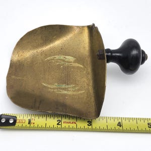 May include: A vintage brass scoop with a dark wooden handle. The scoop is approximately 5 inches long and has a curved shape. The handle is attached with a metal bolt. The brass has a worn, aged appearance.