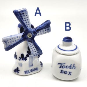 May include: Two ceramic figurines: a windmill and a tooth box. The windmill is white with blue accents and a blue and white patterned windmill. The tooth box is white with a blue lid and the words "Tooth BOX" in blue.