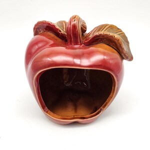 May include: A ceramic apple-shaped container with a glossy red finish. The container has a large opening and is adorned with brown leaves and a stem. The interior is a warm brown color, and the piece is set against a white background.