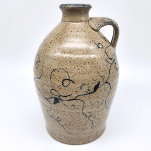 May include: A tan ceramic jug with a handle and a narrow neck. The jug features a speckled surface and is decorated with black, swirling vine-like designs. The handle is arched, and the rim is a darker shade of brown.