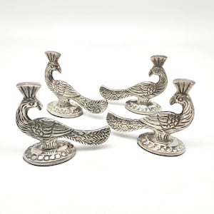 May include: A set of four silver-toned metal peacock figurines, each featuring an upright crest, detailed wing feathers, and a long tail. The birds are mounted on circular, textured bases with a repeating pattern along the outer edge.