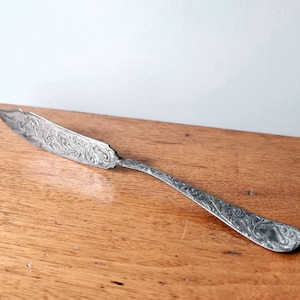 May include: A silver-toned metal butter knife featuring an intricate, embossed scrollwork pattern covering the entire surface of the blade and the handle. The utensil has a curved, pointed tip and a slender, tapered neck leading to a rounded handle.