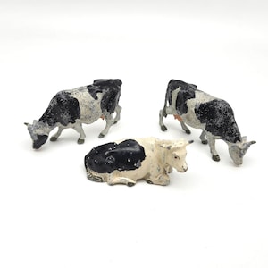 May include: Three small metal cow figurines with a distressed black and white painted finish. Two of the cows are standing in a grazing position with their heads lowered, while the third cow is positioned in the center in a lying down pose.