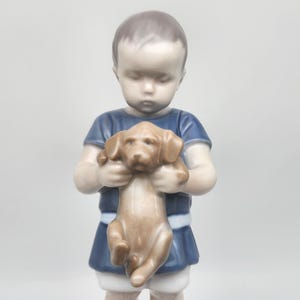 May include: A porcelain figurine of a child in a blue tunic holding a brown puppy. The child has a serene expression, and the puppy is detailed with realistic features. The figurine is set against a plain background.