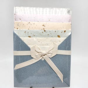 May include: A stationery set featuring a variety of colorful paper sheets, including pink, beige, and blue, with gold flecks. The sheets are held together by a light blue envelope-style holder and tied with a cream-colored ribbon and bow. The set is packaged in clear plastic.