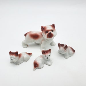 May include: A collection of ceramic bulldog figurines. The set includes a standing bulldog and three smaller bulldogs in a lying down position. The bulldogs are white with brown markings.