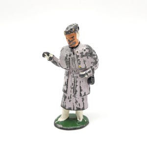 May include: A small, vintage metal figurine of a person in a long coat and hat. The figure is painted in shades of gray, lavender, and black, with a green base. The figure is standing and holding an object in their left hand.