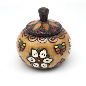 May include: A round, wooden trinket box with a lid. The box is light brown with carved floral designs and colorful painted accents. The lid has a dark brown knob and a floral pattern.