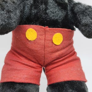 May include: A plush toy featuring black fur and red shorts with two yellow circular buttons. The toy's design is reminiscent of a classic cartoon character, with a focus on the torso and upper legs. The toy is made of soft, plush material.