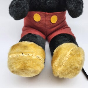 May include: A plush toy of a cartoon character, featuring black fur, red shorts with yellow buttons, and large yellow feet. The toy's design is reminiscent of a classic animated character.