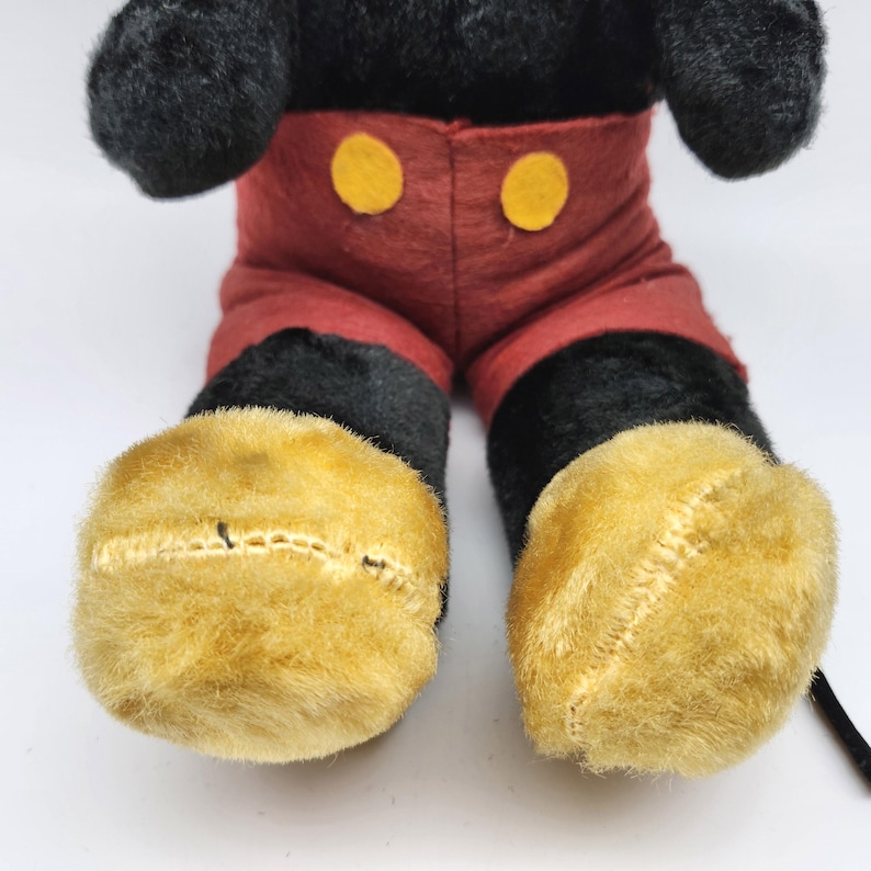 May include: A plush toy of a cartoon character, featuring black fur, red shorts with yellow buttons, and large yellow feet. The toy's design is reminiscent of a classic animated character.