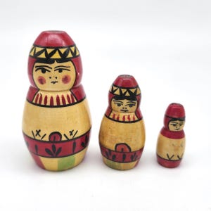 May include: Three wooden nesting dolls, each painted with a traditional design. The dolls are in shades of red, yellow, and green, with black accents. The largest doll is approximately 5 inches tall.