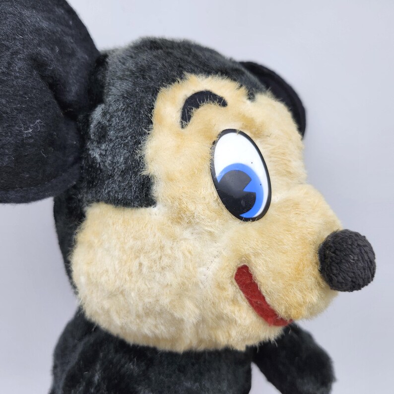 May include: A vintage plush toy mouse with black ears, head, and body. The mouse has a tan face, a black nose, and a red stitched smile. The eyes are large with blue irises and black pupils. The toy is a classic children's item.