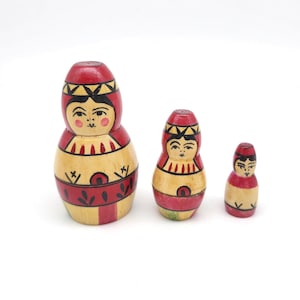 May include: Three wooden nesting dolls, also known as Matryoshka dolls, in varying sizes. The dolls are painted with red, yellow, and black details, featuring a traditional design with faces and floral patterns. The largest doll is approximately 5 inches tall.
