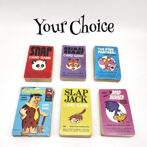 May include: Six vintage card games are displayed, including "Snap," "Animal Rummy," and "The Pink Panther." The games feature colorful illustrations of cartoon characters and are arranged on a white surface. The text "Your Choice" is at the top.