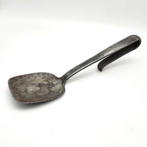 May include: A weathered, dark gray metal spatula with a rectangular, flat head and a long, curved handle. The surface of the utensil shows significant signs of oxidation, wear, and discoloration, giving it a textured, aged appearance throughout.