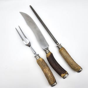 May include: A three-piece carving set consisting of a two-pronged meat fork, a curved carving knife, and a sharpening steel. Each tool features a textured, natural-colored antler handle with silver-toned metal ferrules and end caps.