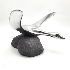 May include: A polished silver metal sculpture of a swan in flight, mounted on two dark gray, textured stone-like forms. The swan's wings are outstretched, and its neck curves gracefully. The sculpture is set against a white background.