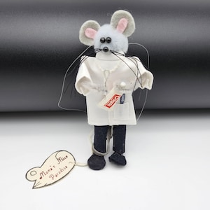 May include: A small gray mouse wearing a white lab coat and black pants, holding a toothbrush and a tube of toothpaste. The mouse has a tag attached to its foot that reads "Mona's Mice Paradise".