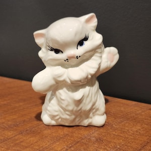 May include: A white ceramic cat figurine with pink accents on the ears and paws. The cat has black eyelashes, blue eyes, and a pink nose. It is posed with one paw raised, giving a friendly wave. The figurine is on a wooden surface.