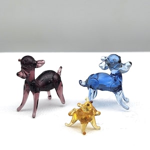 May include: Three miniature glass animal figurines: a purple deer, a blue deer, and a yellow dog. The deer have delicate antlers and slender legs. The dog is small and has a rounded body. All are transparent and appear to be handmade.