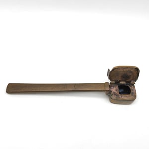 May include: A brass inkwell with a hinged lid and a long, thin handle. The lid is open, revealing a small, rectangular inkwell inside.