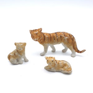 May include: Three ceramic tiger figurines. A large tiger stands with its head turned to the left. Two smaller tigers are lying down. The tigers are painted in shades of brown and white.