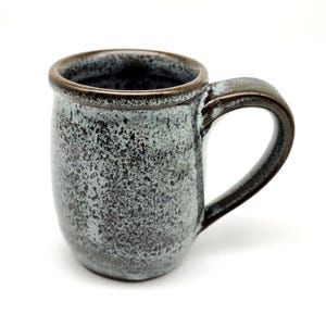 May include: A handmade ceramic mug with a speckled, textured surface in shades of gray and blue. The mug has a dark brown rim and handle. The handle is a rounded, curved shape. The mug is designed for hot beverages.