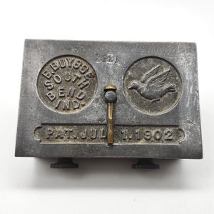 May include: Antique metal stamp with the text "E. Buysse South Bend Ind" in a circle, a dove emblem, and "PAT. JUL 1. 1902". The stamp has a handle and two small knobs. The metal has a worn, aged appearance.