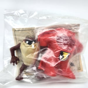 May include: A Taz figure and a red cape with a yellow and black emblem are sealed in a clear plastic bag. The Taz figure is brown, tan, and black. The cape is red. The figure is on a cardboard backing.