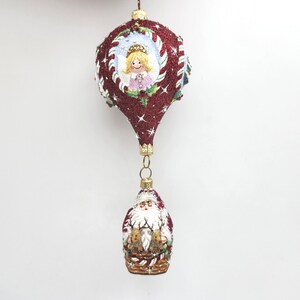 May include: A red and white glass Christmas ornament shaped like a hot air balloon with a painted angel inside. A smaller ornament with Santa Claus and reindeer hangs from the bottom.