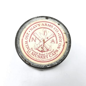May include: A round, vintage metal tin with a red and white label. The label reads "NAVY ARMS CO." and "MUSKET CAPS." The tin has a distressed appearance, with visible wear and tear. The label features crossed cannons and an anchor.