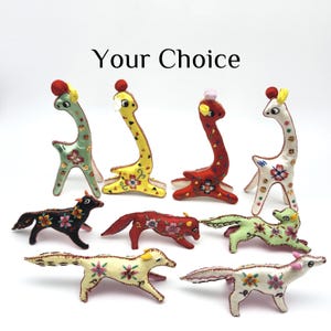 May include: A collection of eight small, handcrafted fabric animal figurines featuring giraffes and foxes. Each figure is decorated with colorful floral embroidery, sequins, and small fabric accents. The text "Your Choice" is centered above the items.