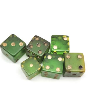 May include: Six green dice with gold-colored dots are arranged on a white surface. The dice are a mix of shades of green, with some having a marbled appearance. Each die has a different number of dots, from one to six.